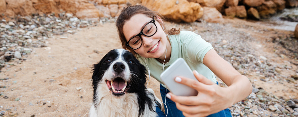 Smiling young cheerful girl in eyeglasses having fun with her dog at the beach and taking selfie with smartphone